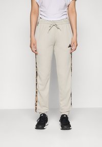 Beige sweatpants med elastisk midje, sider med leopardmønster striper, og avkuttede ankler. Kombinert med svarte sneakers med hvite detaljer.
