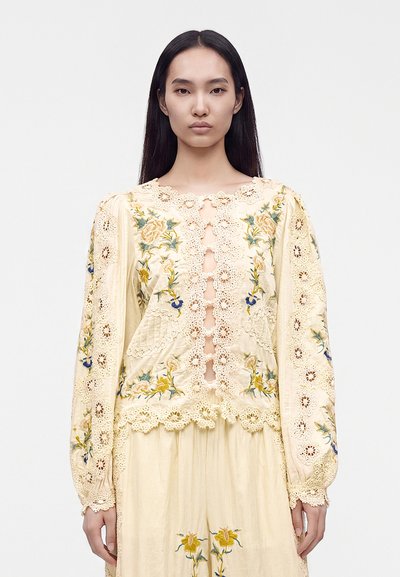 Woman with long straight black hair wearing a cream lace blouse with floral embroidery and matching skirt standing against a plain background.