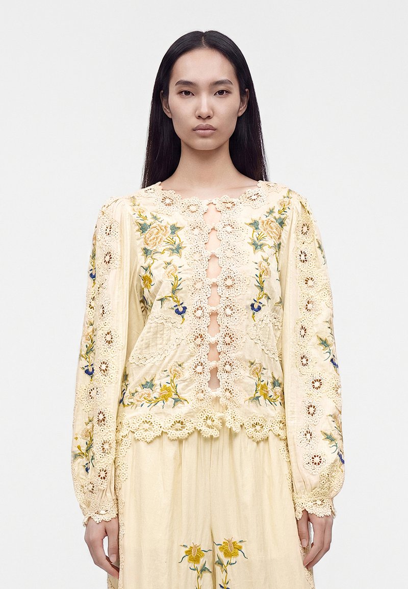 Woman with long straight black hair wearing a cream lace blouse with floral embroidery and matching skirt standing against a plain background.