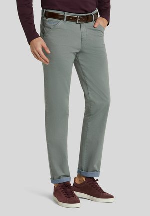 Homme portant un pantalon gris clair retroussé, une ceinture en cuir marron, des baskets bordeaux et une chemise à manches longues violet foncé avec une main dans la poche.