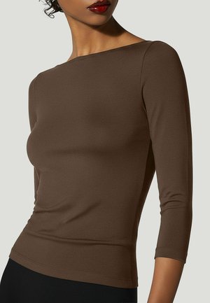 Donna che indossa un top marrone aderente a maniche lunghe con scollo a barca e pantaloni neri, mostrata dal collo ai fianchi su uno sfondo semplice.