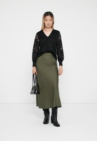 Μαύρη μπλούζα μακρύ μανίκι με δαντελένια επένδυση και V-λαιμόκοψη, συνδυασμένη με μίντι φούστα σε πράσινο χρώμα. Το μοντέλο κρατά μια μικρή μαύρη τσάντα. Μαύρες μπότες μέχρι το γόνατο.