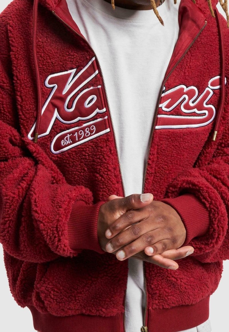 Personne portant une veste zippée rouge texturée avec une inscription blanche "est 1989", mains jointes sur une chemise blanche.