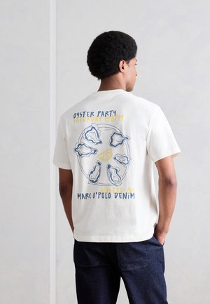 Mand iført hvid T-shirt med østers- og citrongrafik og teksten "Oyster Party Saturday @8PM Hosted by Marco Polo Denim" på ryggen.
