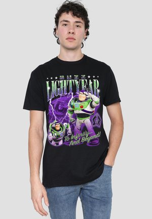Junger Mann trägt ein schwarzes Buzz Lightyear T-Shirt mit grünem und lila Text und Grafiken, kombiniert mit blauen Jeans vor einfarbigem Hintergrund.