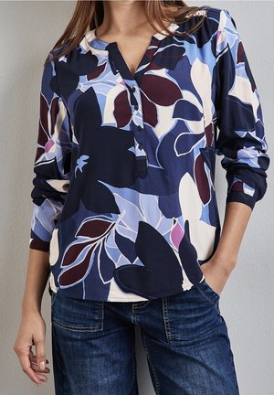 Femme portant un chemisier à manches longues avec de grands motifs floraux bleus, blancs et bordeaux et un jean bleu foncé, debout avec une main dans la poche.