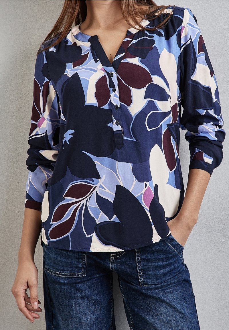 Femme portant un chemisier à manches longues avec de grands motifs floraux bleus, blancs et bordeaux et un jean bleu foncé, debout avec une main dans la poche.