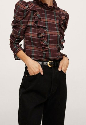 Mujer con blusa de cuadros burdeos con volantes y mangas abullonadas, metida dentro de pantalones negros de tiro alto con cinturón negro.