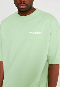 Camiseta de algodón verde claro con mangas cortas. En el frontal se encuentra la palabra "MULTIPLY" en texto blanco en negrita. Textura suave, ajuste casual.