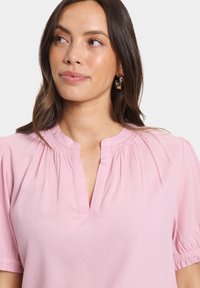 Helles rosa kurzärmliges Blusenshirt mit gerafftem Halsausschnitt, subtiler Textur und einer Knopfleiste. Die Ärmel sind leicht puffig.