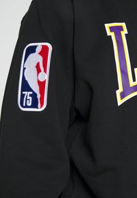 Sudadera negra con el logo de la NBA bordado en la manga izquierda. Diseño audaz de una "L" morada en la parte frontal. Tejido suave con una textura lisa.