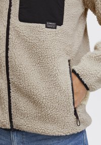 Beige fleece jacket with textured surface, black chest pocket, and zipper details. Features elastic cuffs and a casual fit.