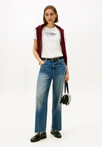 Hvid T-shirt med sort logo, parret med brede blå denimjeans med frynsede kanter. Burgunder vest hængende over skuldrene, sort håndtaske.