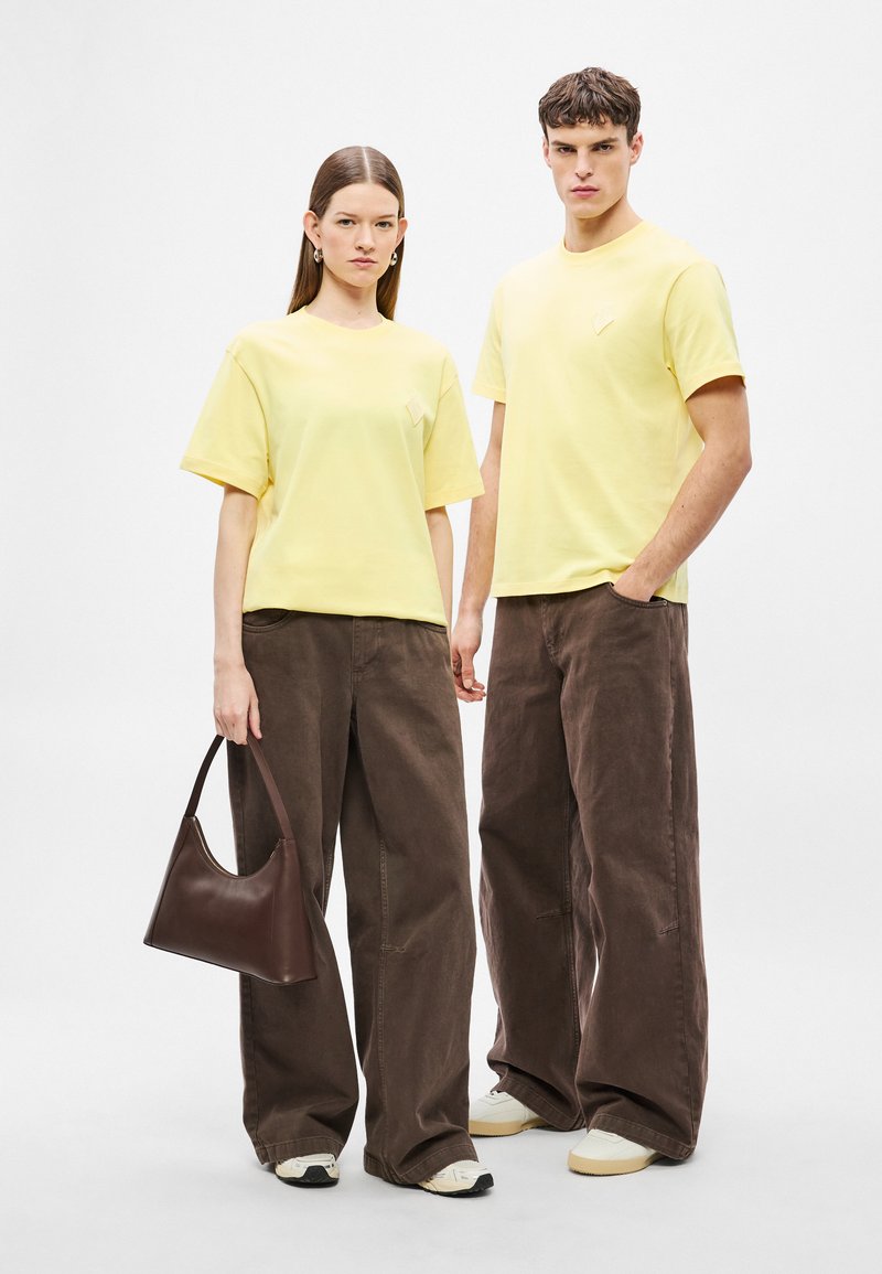 T-shirt gialle in cotone con scollo rotondo, pantaloni marroni a gamba larga in velluto a coste. Accessori includono una borsa in pelle marrone e sneakers bianche.