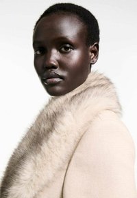 Beige coat with a soft, light fur collar. The fabric is smooth and tailored, highlighting clean lines and a structured silhouette.