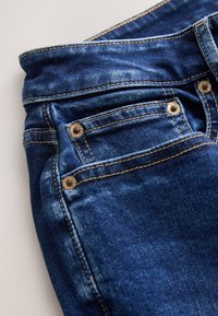 Gros plan sur un jean en denim bleu foncé plié montrant la poche avant, la poche-monnaie avec rivets métalliques, et les coutures visibles sur un fond clair.