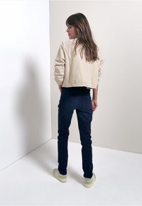 Chaqueta corta beige sobre una camiseta negra, combinada con jeans oscuros de corte ajustado y zapatillas de color claro. Tejido suave con textura sutil.