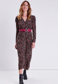 Abito lungo floreale scuro con maniche lunghe e scollatura a V profonda. Accentuato da una larga cintura rosa e indossato con stivaletti neri. Tessuto liscio.