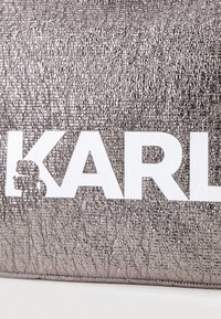 Metallic sidabrinės spalvos krepšys su tekstūruota paviršiumi, kuriame išryškintas baltas "KARL" logotipas ir grafiška silueto iliustracija vienoje pusėje.