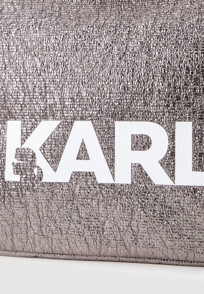 Metallic sidabrinės spalvos krepšys su tekstūruota paviršiumi, kuriame išryškintas baltas "KARL" logotipas ir grafiška silueto iliustracija vienoje pusėje.