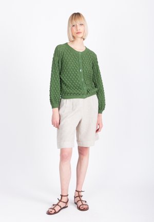 Mujer con peinado bob rubio vistiendo un cárdigan verde texturizado, pantalones cortos beige hasta la rodilla y sandalias marrones con tiras, de pie contra un fondo blanco.