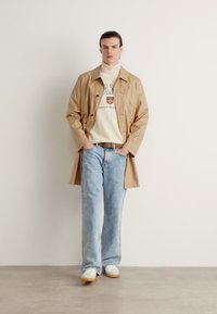 Trench coat impermeabilizat bej peste un pulover alb cu gât înalt, combinat cu blugi largi de culoare albastru deschis și adidași albi. Texturi netede în întregime.