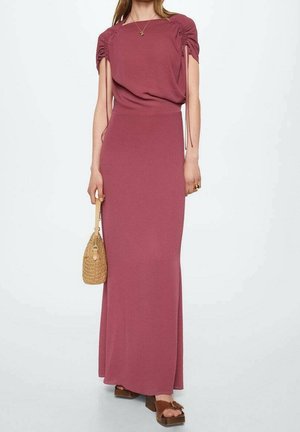 Frau trägt ein mauvefarbenes, gerafftes Maxikleid mit kurzen Ärmeln, hält eine beige geflochtene Handtasche und trägt braune Plateausandalen.
