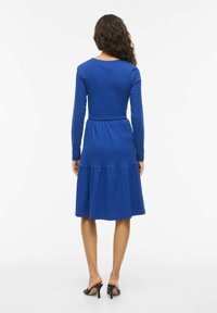 Modré bavlněné šaty s dlouhým rukávem, s vícedílnou sukní, kulatým výstřihem a elastickým pasem, doplněné černými botami na podpatku typu mule.