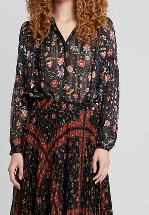 Vrouw draagt een doorschijnende zwarte blouse en geplooide rok met rode en bloemmotieven, staand tegen een effen lichtgrijze achtergrond.