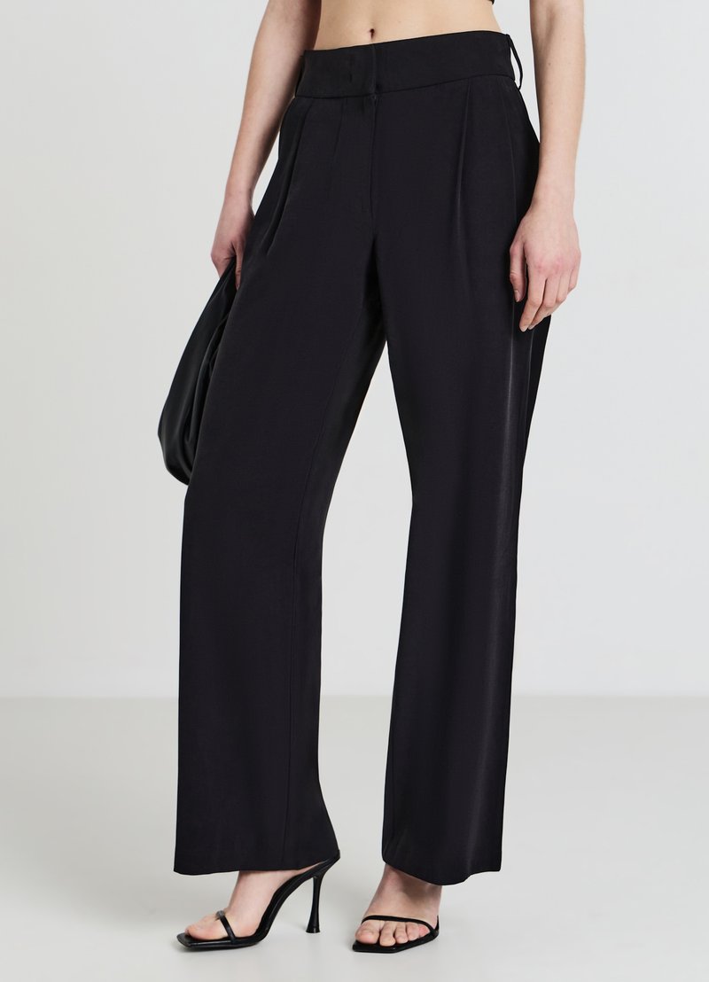Schwarze weit geschnittene Hose mit Falten, ausgestattet mit einer glatten Textur und hohem Taillenbund. Kombiniert mit schwarzen Sandalen mit Riemchen und einer schwarzen Tasche.