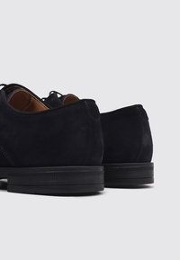 Chaussures en daim bleu marine avec une finition texturée, dotées d'un profil bas, d'une forme arrondie et de semelles en caoutchouc noir. Les lacets passent par des œillets.
