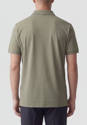 Uomo che indossa una polo verde oliva a maniche corte e pantaloni scuri, visto da dietro contro uno sfondo chiaro e uniforme.