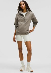 Lys brun zip-up sweatshirt med høj krave, sidelommer, ribbede manchetter, parret med cremefarvede cykelshorts og lyse sneakers.