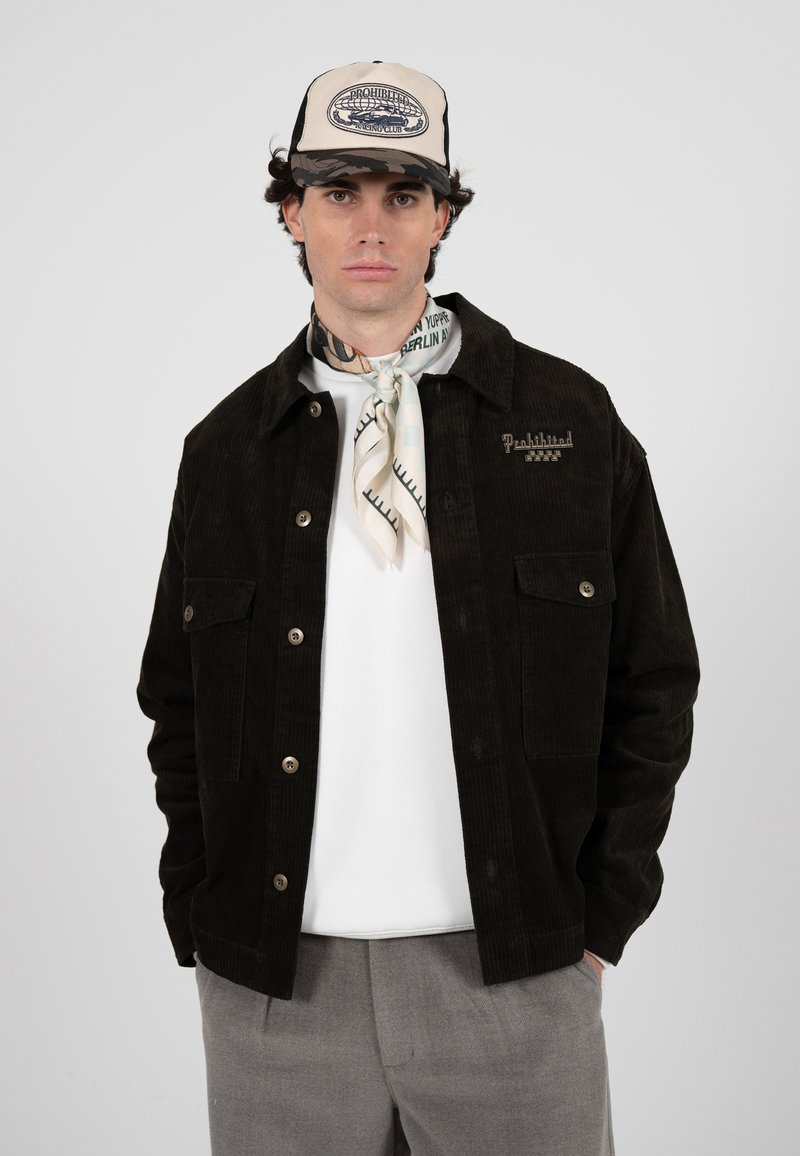 Junger Mann, der eine schwarze Cordjacke, ein weißes Hemd, ein Halstuch, graue Hose und eine beige Baseballkappe mit Camouflage-Muster vor einem einfarbigen Hintergrund trägt.