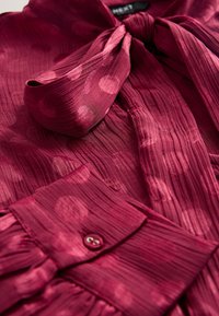 Blusa color amaranto con tessuto testurizzato, caratterizzata da grandi motivi circolari. Comprende un collo annodato e dettagli con polsini abbottonati. Design morbido e fluido.