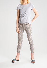 Grå t-shirt med kort ärm, kamouflage-mönstrade smala cargobyxor i beige toner och svarta slip-on sandaler med gröna och vita accenter.