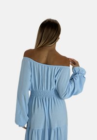 Robe bleu clair, à épaules dénudées, avec des manches longues, taille smockée et jupe à volants. Texture douce, design fluide.