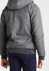 Grå huvtröja med en texturerad yta, elastiska ärmslut och nederdel. Har en svart lager av stickad trim vid midjan. Enkel, funktionell design.