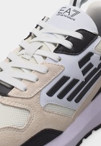 Λευκό και μπεζ αθλητικό παπούτσι με μαύρες λεπτομέρειες, λευκά κορδόνια, υφή από δίχτυ και σουέντ, και το λογότυπο EA7 Emporio Armani στη γλώσσα και στο πλάι.