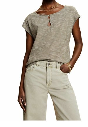 Kvinde iført lyse beige højttaljede bukser og en kortærmet taupe stribet top med nøglehulsknapper, tilbehør med guldsmykker.