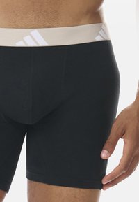 Boxer shorts noirs avec une ceinture beige arborant un logo blanc. Le tissu est lisse avec une coupe ajustée et des coutures minimales.