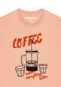 Perzikkleurig T-shirt met een zwarte omtrek van een French press en twee glazen, met rode tekst die zegt: "KOFFIE maakt alles beter."