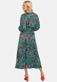 Bunt gemustertes Maxikleid mit lebhaftem Blumenmuster, tailliertem Schnitt, langen Ärmeln und fließender Silhouette. Kombiniert mit klobigen roten Absätzen.