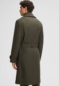 Manteau en laine vert olive avec une taille ceinturée, de grands revers et des poches latérales. Le tissu a une finition texturée et une coupe mi-longue.