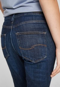 Dark blue denim jeans with a fitted design. Features orange stitching and two back pockets with a curved decorative seam. Smooth texture.