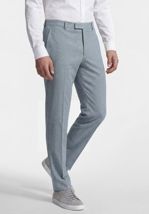 Man draagt lichtblauwe op maat gemaakte broek, wit geknoopt overhemd en grijze sneakers, staand tegen een effen achtergrond.