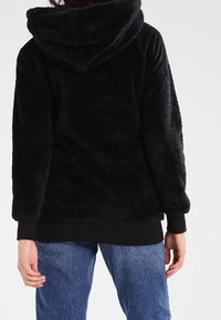 Musta fleecehuppari, jossa pehmeä pinta, huppu ja resorit hihansuissa, yhdistetty sinisiin denimhousuihin. Ei näkyviä logoja tai kuvioita.