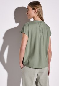 Blusa verde a maniche corte con una superficie liscia, dettaglio plissettato sulla parte alta della schiena, abbinata a pantaloni grigi chiari.
