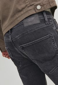 Muškarac u crnim trapericama s vidljivim stražnjim džepovima i smeškim jaknom, ruka mu je naslonjena na bok, fokus na donji deo torza.