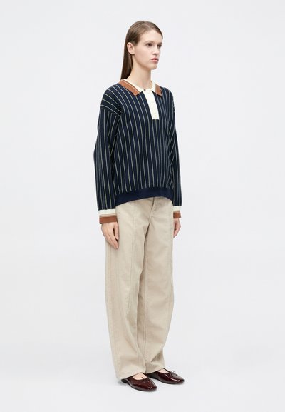 Πλεκτό πουλόβερ με ναυτικό μπλε και κρεμ ρίγες, με καφέ γιακά, συνδυασμένο με μπεζ παντελόνι φαρδιάς γραμμής και σκούρα κόκκινα παπούτσια. Απλός, μοντέρνος σχεδιασμός.